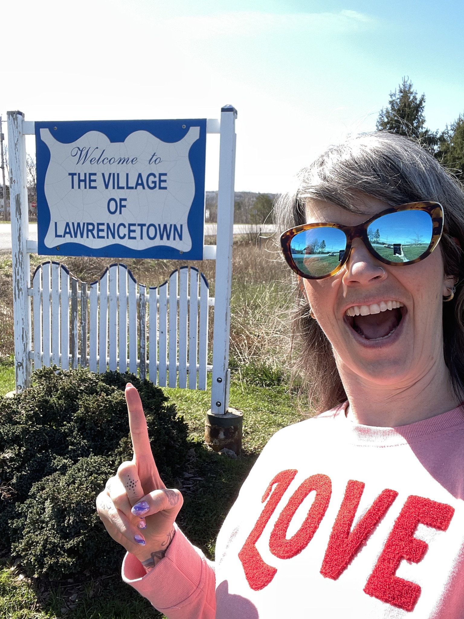 The town sign for the Village of Lawrencetown is in the back ground. A woman wearing a pink sweater with the word "love" in red is pointing to the sign with an open-mouth, very excited smile.