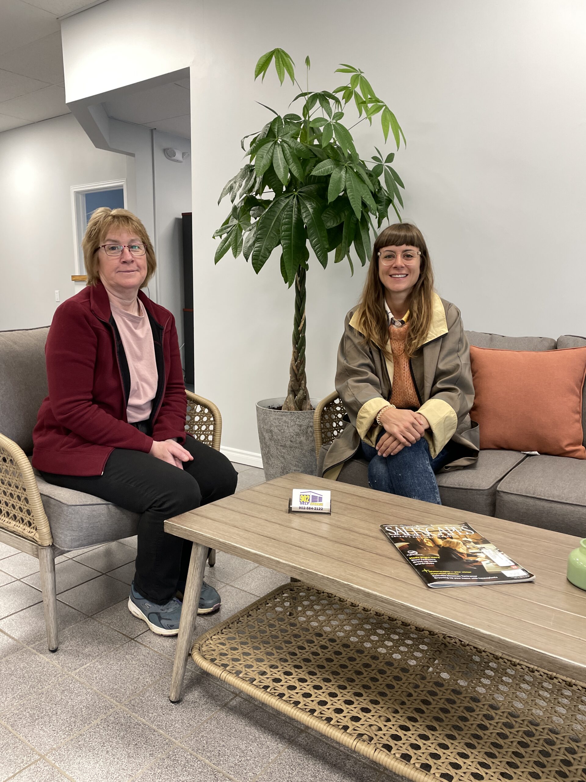 Two women sit in a bright lounge or lobby area. They are facing the camera and smiling.