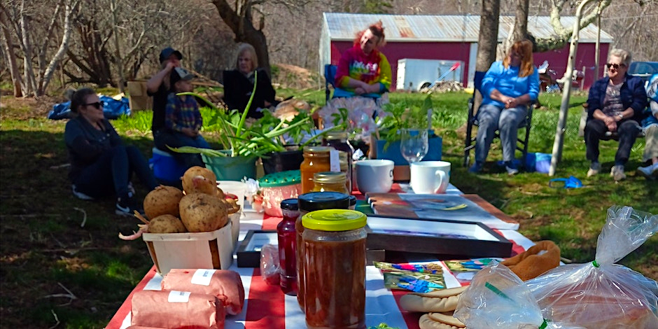 A table full of homemade and homegrown food is in the foreground. A group of people sitting in lawn chairs on grass are in the background.