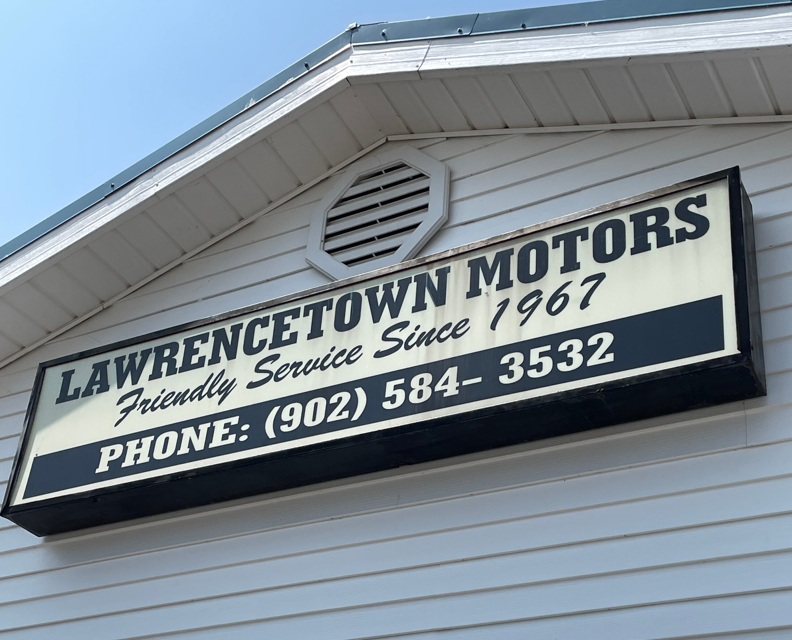 A white sign with blue text on the side of a white building reads "Lawrencetown Motors, Friendly Service since 1967. Phone 902-584-3532"